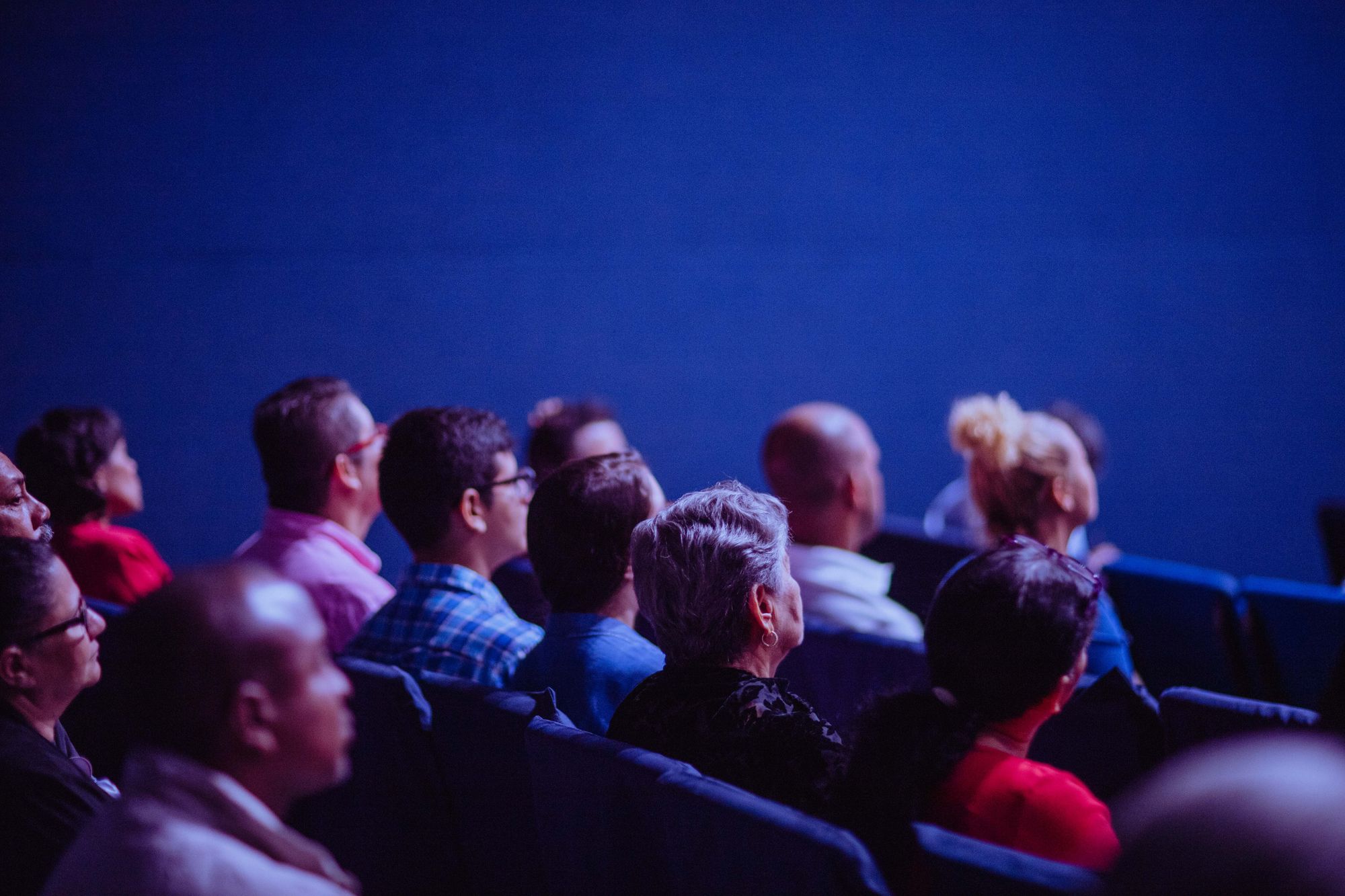 Rows of conference delegates in a darkened auditorium, watching a presentation off-frame