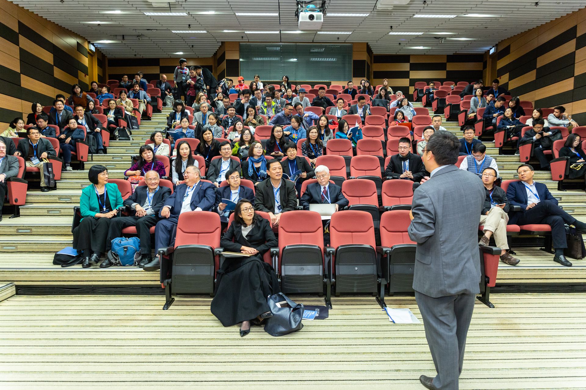A speaker standing at the front of a tiered lecture hall, addressing a seated professional audience wearing conference lanyards