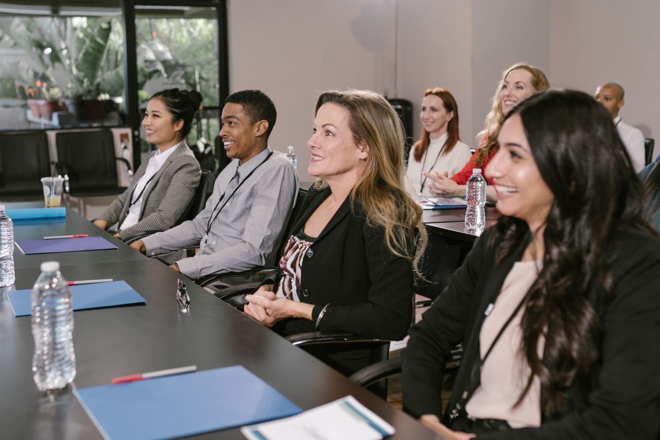 Six business attendees seated along a conference table at a session, listening and smiling, wearing lanyards