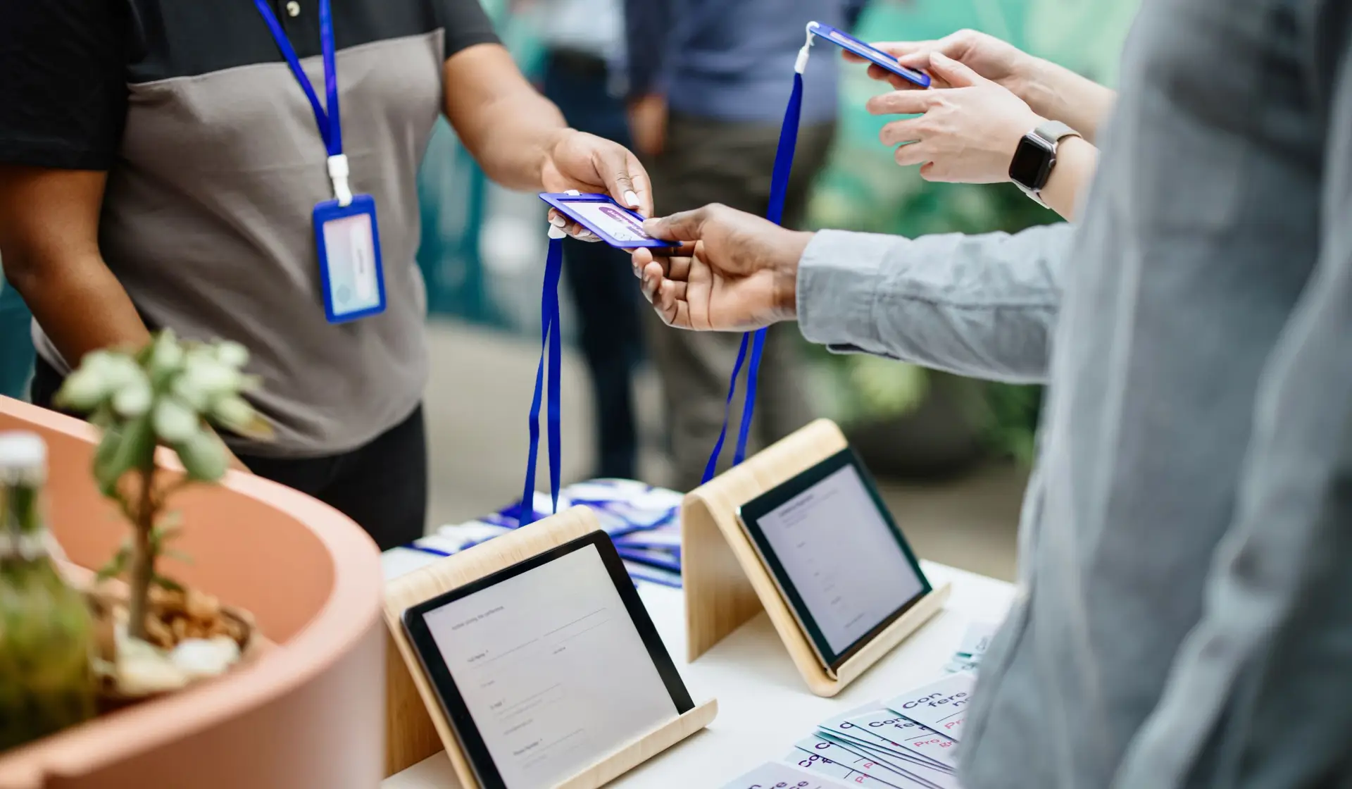 A staff member passing a printed lanyard badge across a registration counter to an attendee, with tablets on stands and spare lanyards on the desk